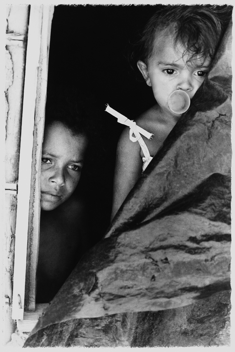 Foto em preto e branco de duas crianças olhando pela janela. A menor, na parte superior direita, apoiada no que parece ser uma lona plástica, tem algo na boca que lembra uma chupeta. Ambas estão com olhar vago