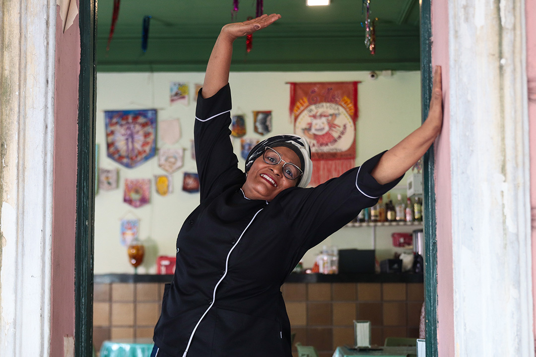 Dona Rosa de braços levantados, sorridente, em uma das portas de entrada do Hotel