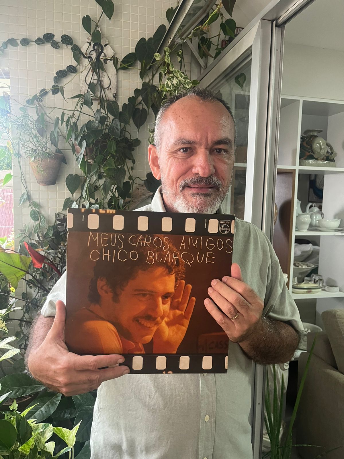 O empresário segurando o disco de Chico Buarque na varanda de uma casa ou apartamento com várias plantas ao fundo