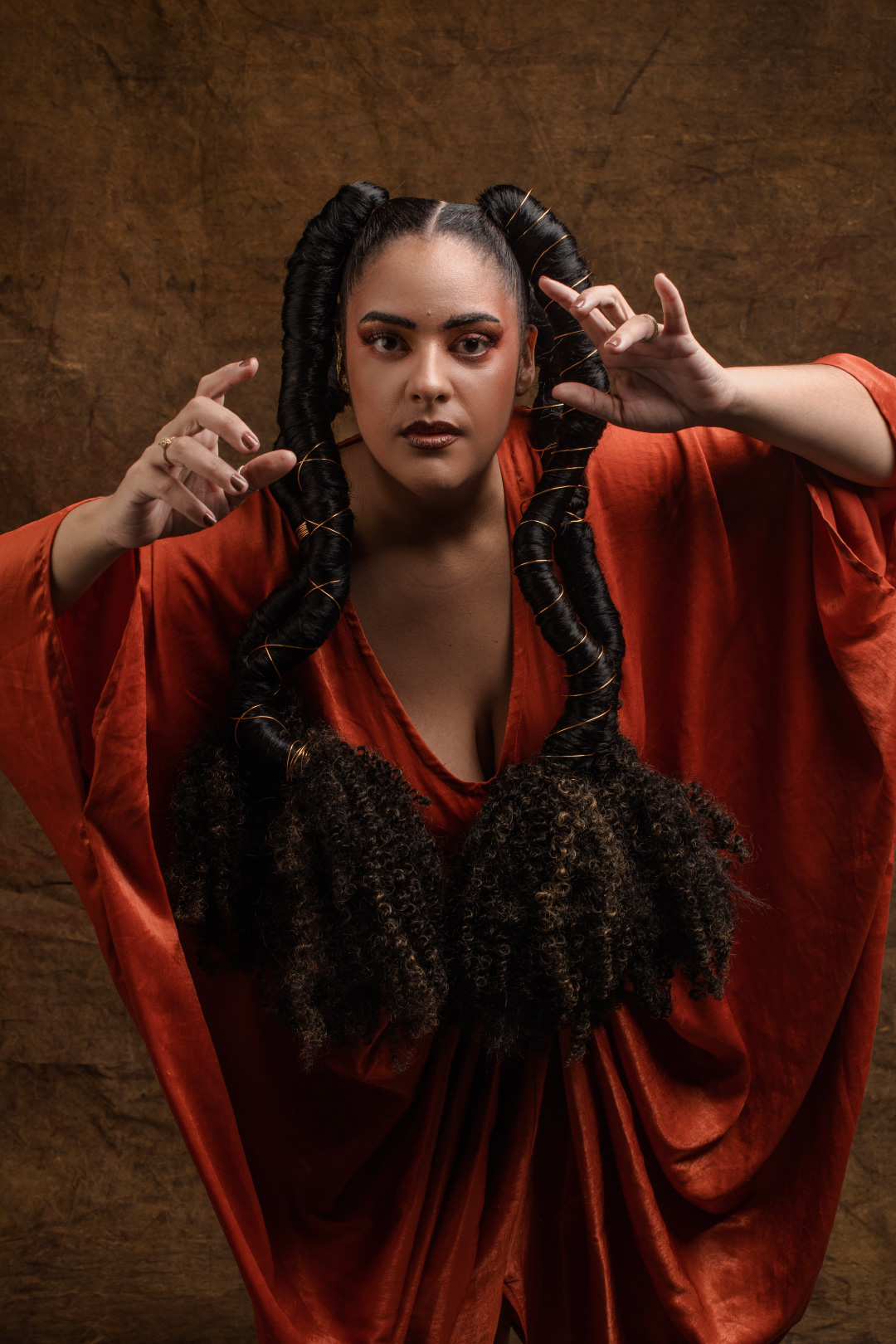 A imagem mostra uma mulher posando dramaticamente diante de um fundo marrom texturizado, em um estúdio fotográfico com iluminação baixa e cinematográfica. Ela veste uma roupa ampla e fluida em tom vermelho-alaranjado, com mangas largas que lembram um figurino teatral ou ritualístico. O cabelo é o elemento mais marcante da composição: dividido ao meio, forma duas estruturas altas e enroladas no topo da cabeça, descendo em longos cachos volumosos até a altura do peito. O penteado cria uma silhueta forte e quase escultórica. Ela usa argolas douradas grandes e maquiagem intensa, com destaque para os olhos e o batom escuro. A modelo encara diretamente a câmera com expressão séria e imponente. As mãos estão erguidas em direção ao observador, com os dedos curvados, como se estivesse lançando um feitiço, conduzindo uma performance ou incorporando uma personagem mística. A iluminação destaca o rosto e o tecido avermelhado, enquanto o restante da cena permanece em sombras suaves, reforçando o clima dramático e sobrenatural da fotografia.